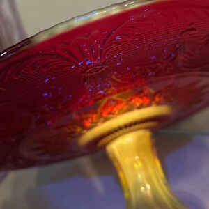 May include: A red glass pedestal cake stand with a decorative floral pattern. The stand has a yellow stem and a scalloped edge. The glass has an iridescent quality, reflecting light with a rainbow effect.
