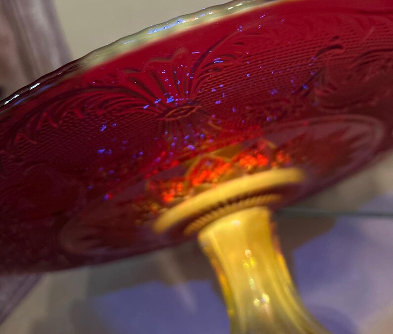 May include: A red glass pedestal cake stand with a decorative floral pattern. The stand has a yellow stem and a scalloped edge. The glass has an iridescent quality, reflecting light with a rainbow effect.
