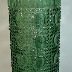 May include: A green glass bottle with a textured, geometric pattern. The bottle has a wide mouth and a rounded bottom.