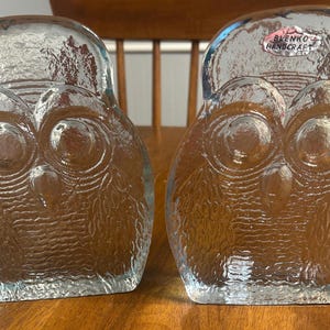 Puede incluir: Dos figuras de búho de cristal transparente con detalles texturizados. Cada búho tiene ojos grandes y redondos y un pico. Las figuras se exhiben sobre una superficie de madera. Una figura tiene una etiqueta que dice "BLENKO HANDCRAFT".