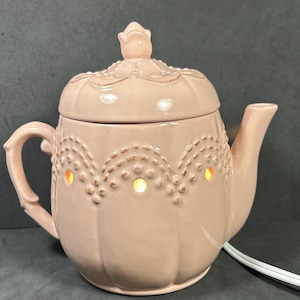 May include: A pink ceramic teapot-shaped wax warmer with a lid and handle. The warmer has a decorative dotted pattern and emits a warm glow through the openings. A white power cord is attached.