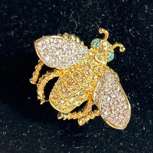 Op de afbeelding: Een gouden bij broche met heldere en lichtblauwe strass steentjes. De bij heeft een gedetailleerd lichaam en vleugels.