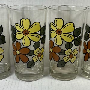 May include: A set of four clear glass tumblers, each featuring a repeating floral pattern. The design consists of large yellow flowers, smaller brown flowers, and dark olive green leaves printed on the exterior surface of the cylindrical glasses.