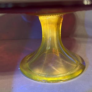 May include: A clear glass cake stand with a yellow stem and a red-purple top. The stem has a flared base and a detailed, ribbed design. The stand is likely used for displaying desserts or other food items.