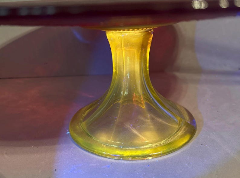 May include: A clear glass cake stand with a yellow stem and a red-purple top. The stem has a flared base and a detailed, ribbed design. The stand is likely used for displaying desserts or other food items.