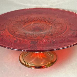 May include: A ruby red glass cake stand with a round, ornate top and a pedestal base. The top features a detailed floral and geometric pattern. The stand is approximately 10 inches in diameter.