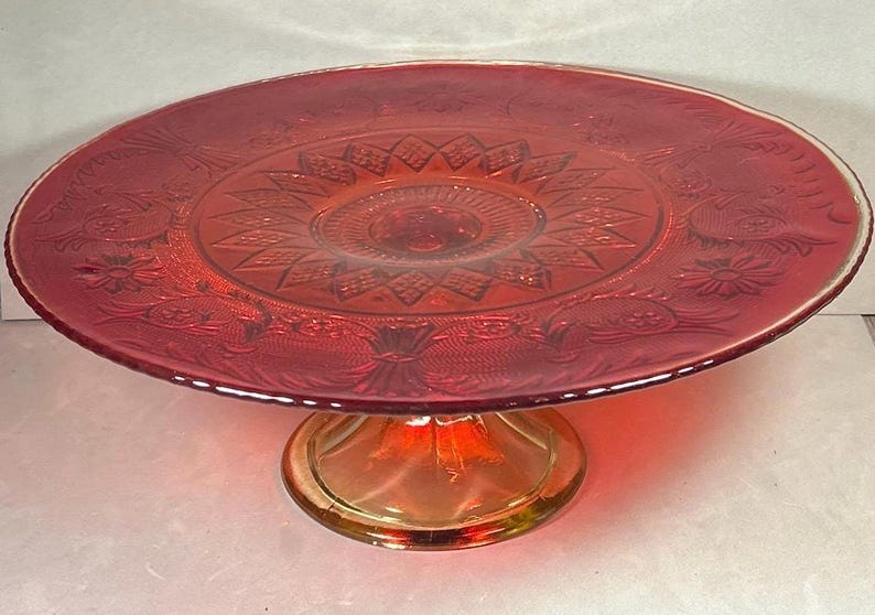 May include: A ruby red glass cake stand with a round, ornate top and a pedestal base. The top features a detailed floral and geometric pattern. The stand is approximately 10 inches in diameter.