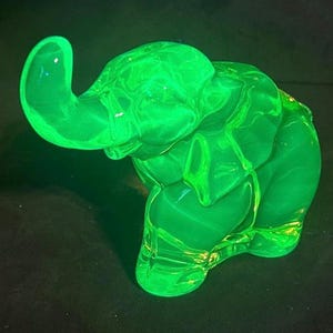 May include: A vibrant, neon green glass elephant figurine. The elephant is sculpted with detailed features, including a raised trunk and visible wrinkles. The translucent material allows light to pass through, creating a glowing effect. The figurine is a decorative item.