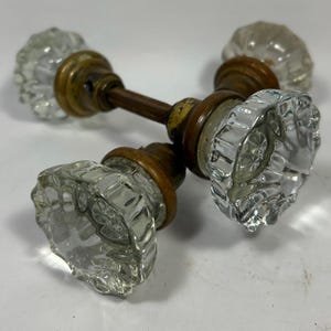 Op de afbeelding: Een set van twee heldere, gefacetteerde glazen deurknoppen verbonden door een centrale metalen stang. Elke knop heeft een cirkelvormig, gegroefd ontwerp met een reflecterend oppervlak, bevestigd op een doffe, messingkleurige metalen basis.