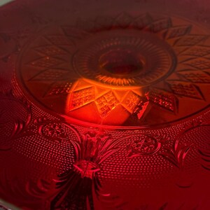 May include: Close-up of a red glass cake stand with intricate embossed floral and geometric patterns. The stand has a central raised portion and a decorative rim. The glass reflects light, creating a warm, inviting glow. A vintage-style serving piece.