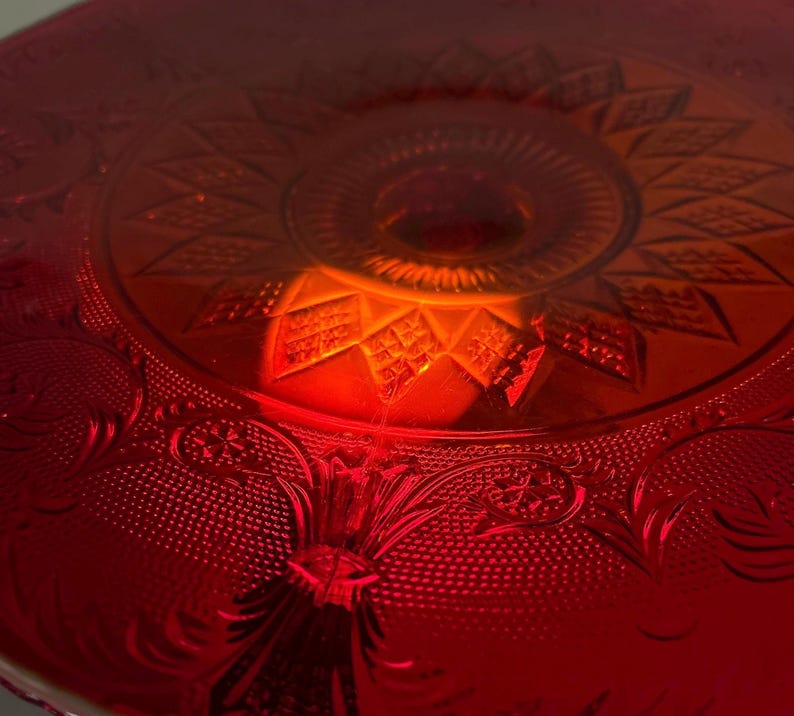 May include: Close-up of a red glass cake stand with intricate embossed floral and geometric patterns. The stand has a central raised portion and a decorative rim. The glass reflects light, creating a warm, inviting glow. A vintage-style serving piece.