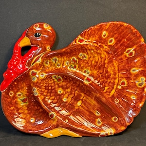 Puede incluir: Una fuente de cerámica con forma de pavo y acabado brillante. La fuente presenta tonos marrones, naranjas y amarillos, con detalles rojos en la carúncula y un ojo negro. Las plumas de la cola del pavo están extendidas.