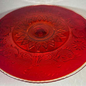 May include: A vibrant red glass cake stand with intricate embossed floral and geometric patterns. The stand features a central pedestal and a wide, scalloped rim. The glass has a glossy finish and a slight iridescent sheen.