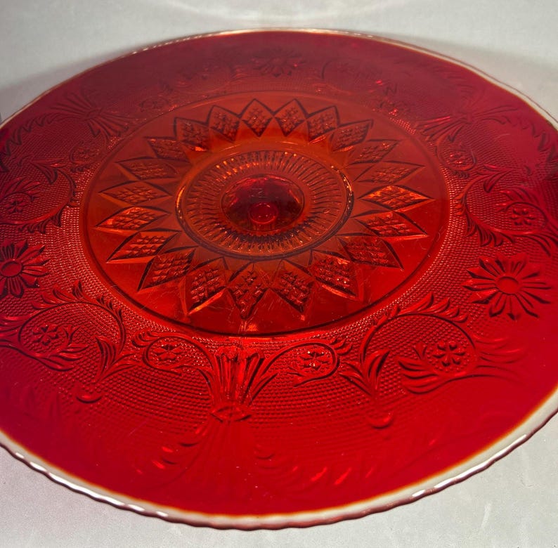 May include: A vibrant red glass cake stand with intricate embossed floral and geometric patterns. The stand features a central pedestal and a wide, scalloped rim. The glass has a glossy finish and a slight iridescent sheen.