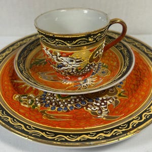 May include: A porcelain tea cup, saucer, and plate set featuring a vibrant orange background with raised white and gold dragon motifs. The edges are decorated with a black border containing gold wave patterns. The interior of the cup is white.