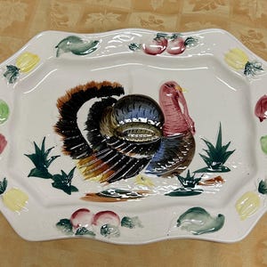 May include: A white ceramic serving platter with a scalloped edge, featuring a hand-painted turkey in the center. The turkey has a colorful plumage and a red head. The border is decorated with painted fruits and vegetables.