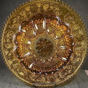 May include: Amber-colored glass serving plate with a detailed floral and starburst pattern. The plate has a scalloped edge and a central star design. The glass has a textured surface and is likely vintage.