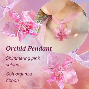 Op de afbeelding: Een glinsterende roze orchideehanger met een zacht organzalint. De hanger heeft de vorm van een bloem. De tekst "Orchid Pendant" is zichtbaar in de afbeelding.