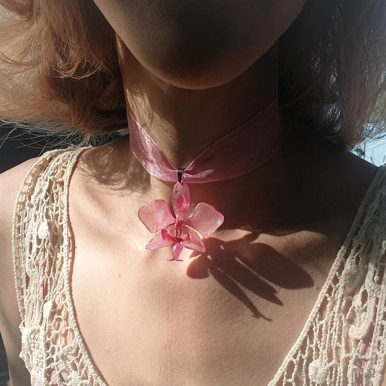 Op de afbeelding: Een roze choker-ketting met een delicate orchideehanger. De orchidee heeft een lichtroze kleur met ingewikkelde details. De ketting wordt om de nek gedragen.