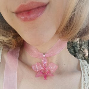 Op de afbeelding: Een roze orchidee hanger ketting. De hanger is een doorschijnende roze bloem met een zilverkleurige metalen sluiting. De ketting is een doorschijnend roze lint. De ketting is een delicaat sieraad.
