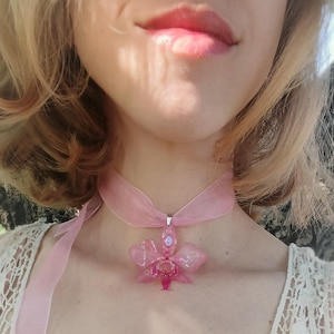 Op de afbeelding: Een roze choker ketting met een doorschijnende orchidee hanger. De hanger heeft een gedetailleerd bloemenontwerp met een centrale edelsteen. De choker is gemaakt van een doorschijnend roze lint.