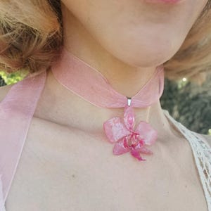 Op de afbeelding: Een roze choker-ketting met een doorschijnende orchideehanger. De orchidee heeft een gedetailleerd, gelaagd ontwerp met een donkerder roze centrum. Het lint is een zacht, transparant materiaal.