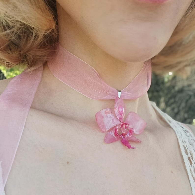 Op de afbeelding: Een roze choker-ketting met een doorschijnende orchideehanger. De orchidee heeft een gedetailleerd, gelaagd ontwerp met een donkerder roze centrum. Het lint is een zacht, transparant materiaal.
