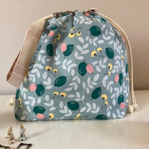 Peut inclure: Un sac bleu clair à cordon de serrage avec un motif botanique de fruits verts et roses et de feuilles blanches. Le sac a une sangle beige et un cordon de serrage blanc. Accessoires et fil visibles.
