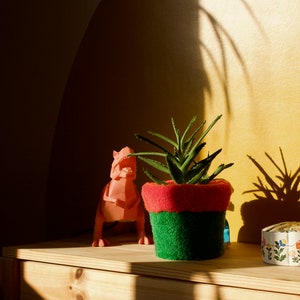 Puede incluir: Una figurita de dinosaurio rosa, una planta suculenta en maceta con hojas verdes y una caja decorativa blanca con diseños florales se muestran en un estante de madera.