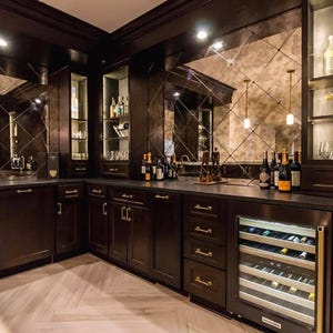 May include: A dark wood home bar with a mirrored backsplash and built-in cabinets. The bar features a wine cooler, bottles of wine, and glassware. The floor has a light-colored herringbone pattern.