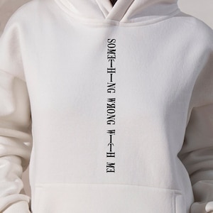Op de afbeelding: Witte hoodie met de tekst "EM-TH-H-I-NG WRONG W-TH-H ME" verticaal in het midden gedrukt.