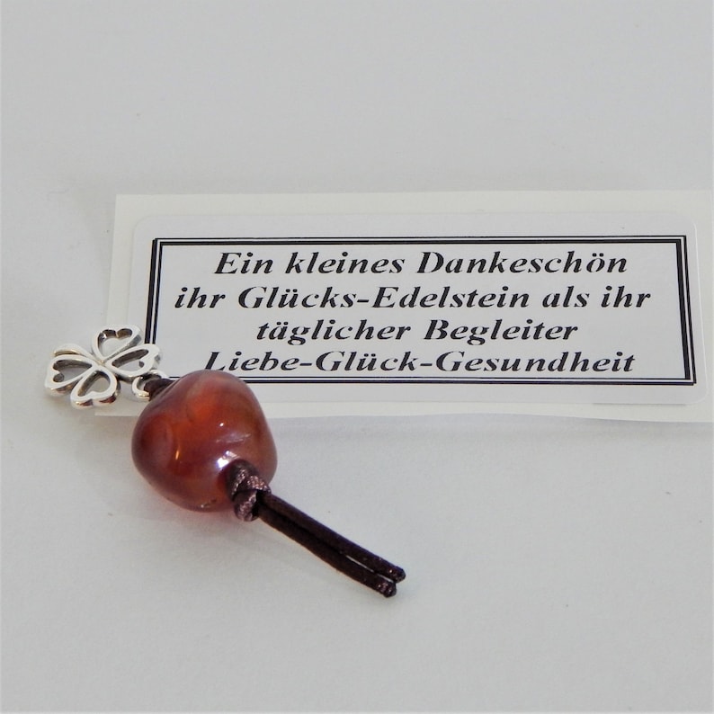 May include: A small brown stone with a silver four-leaf clover charm attached to a brown cord. The stone is a lucky charm and the card reads "Ein kleines Dankesch&ouml;n ihr Gl&uuml;cks-Edelstein als ihr t&auml;glicher Begleiter Liebe-Gl&uuml;ck-Gesundheit".
