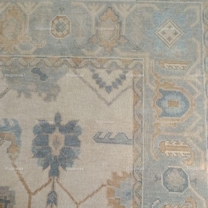 May include: Close-up of a beige and blue patterned rug. The rug features a floral design with blue and tan accents. The border has a geometric pattern in shades of blue and beige. The rug has a fringe edge.