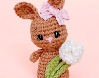 May include: A crocheted brown bunny with a pink bow on its head, holding a white and green crocheted flower. The bunny has black eyes and pink accents. The background is a solid light pink.
