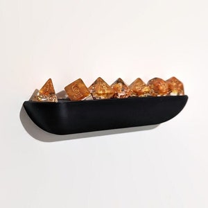 May include: A black, curved, wall-mounted shelf holds a collection of amber-colored dice with gold flecks. The dice are in a variety of shapes and sizes.