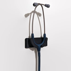Puede incluir: Un estetoscopio azul colgado de un soporte de pared negro. El estetoscopio tiene un tubo plateado y auriculares grises.