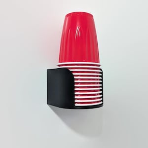 May include: A black plastic cup dispenser mounted on a white wall. The dispenser holds a stack of red plastic cups with white rims.