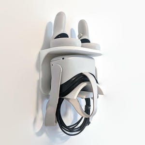 May include: White wall mount for a virtual reality headset with two controllers. The headset is hanging on the mount with the controllers on the top shelf. The headset is white with black straps and a black cable.