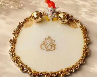 Può includere: Un vassoio rotondo in resina bianca con bordo in foglia d'oro testurizzato, caratterizzato da un simbolo Ganesha dorato centrale. Il vassoio include due piccole ciotole in metallo dorato e una statuetta tridimensionale di Ganesha con turbante.
