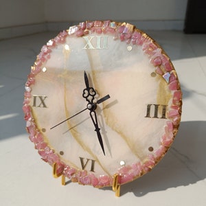 Könnte beinhalten: Eine runde Uhr mit einem weiß-gold marmorierten Zifferblatt, schwarzen Zeigern und goldenen römischen Ziffern. Der äußere Rand ist mit kleinen, grob geschliffenen rosa Steinen verziert. Die Uhr steht auf einem goldfarbenen Metallständer.