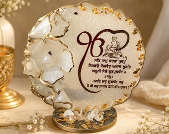 May include: A circular resin plaque featuring gold leaf flakes, white floral accents, and a pearl. The surface displays the Ik Onkar symbol, an illustration of Guru Nanak Dev Ji, and Gurmukhi script text in dark brown, mounted on a round base.
