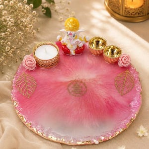 Round pink and white resin pooja thali with a jagged gold-flecked edge, featuring a  Ganesha figurine, a white tealight candle in a gold holder, two small gold diyas, pink rose accents, and gold leaf decorations, with a small circular Gayatri Mantra inscription etched in the center.