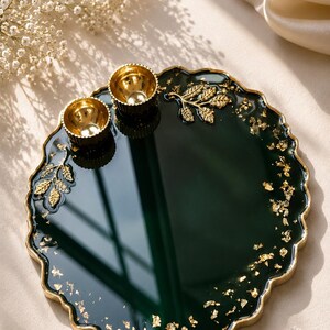 Round dark green resin pooja thali with a wavy gold-trimmed edge, gold leaf sprig accents, scattered gold flecks, and two small gold diyas at the top.