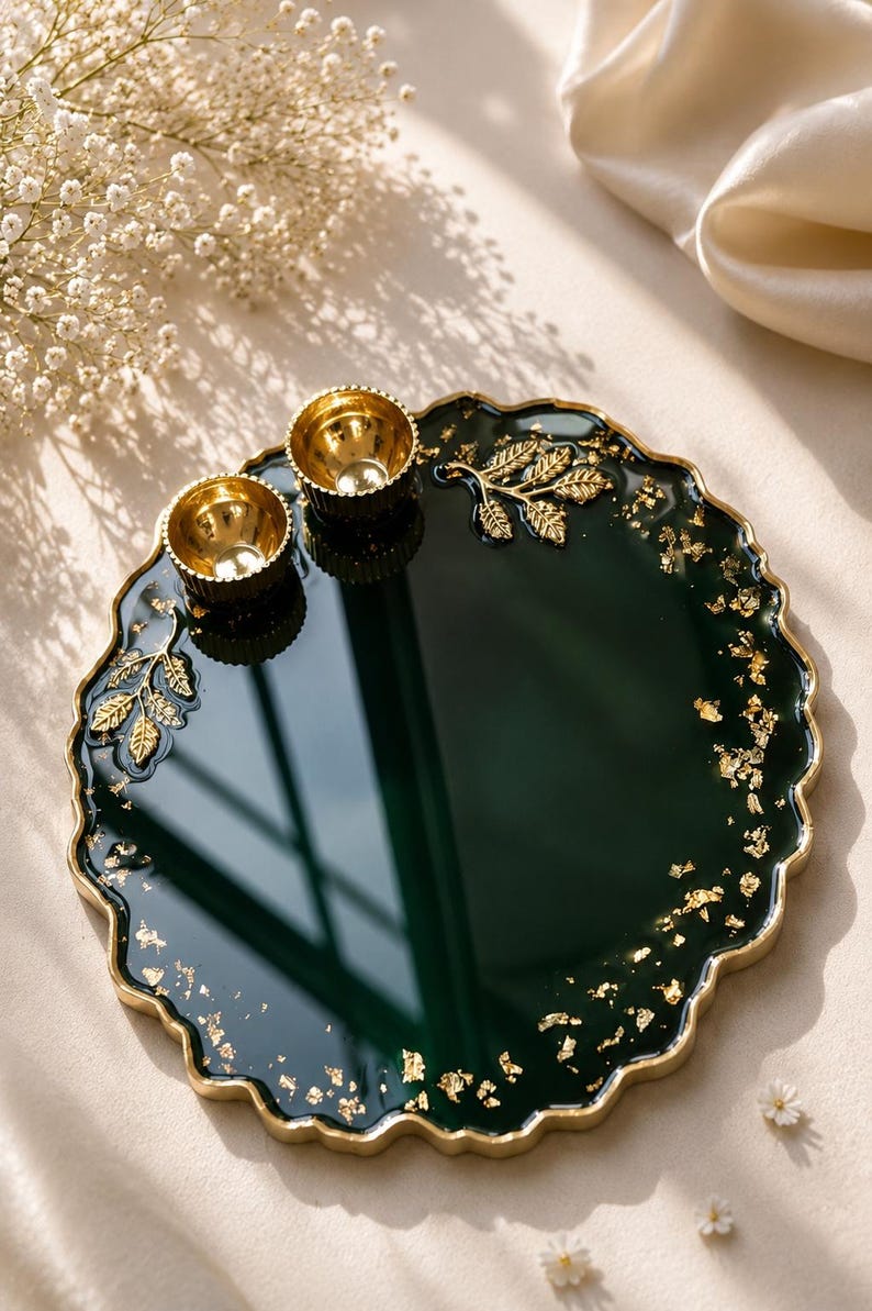 Round dark green resin pooja thali with a wavy gold-trimmed edge, gold leaf sprig accents, scattered gold flecks, and two small gold diyas at the top.