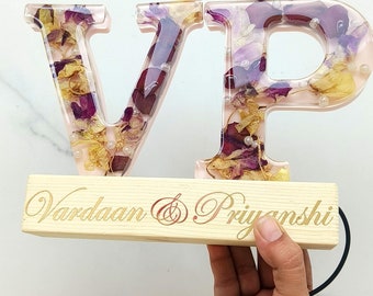May include: A light pink resin letter V and P containing dried purple and yellow flower petals and small white beads. The letters are mounted on a rectangular light wood base engraved with the text Vardaan & Priyanshi and a black power cord.