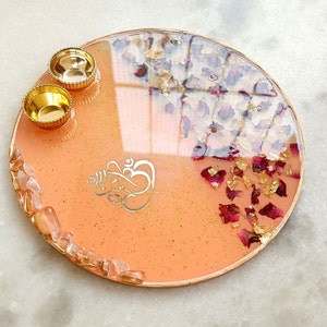 A circular resin tray in peach with gold glitter, featuring a gold Ganesha symbol, two small gold bowls, embedded dried red rose petals, gold leaf flakes, and a border of clear quartz-like stones with a metallic gold rim.
