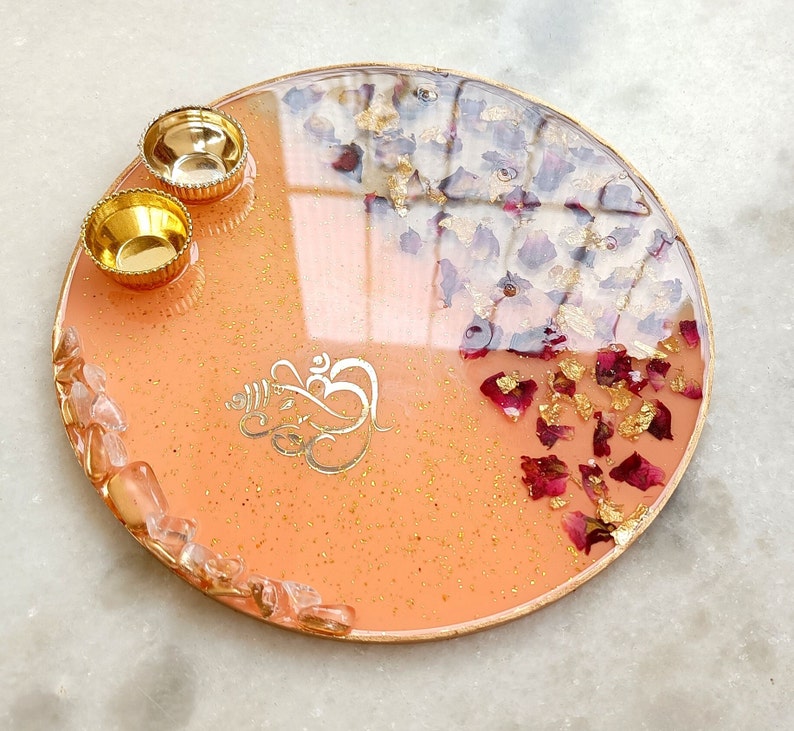 A circular resin tray in peach with gold glitter, featuring a gold Ganesha symbol, two small gold bowls, embedded dried red rose petals, gold leaf flakes, and a border of clear quartz-like stones with a metallic gold rim.