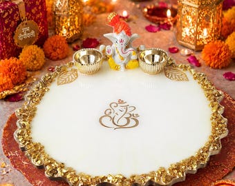 Può includere: Un vassoio decorativo circolare bianco con un emblema dorato di Ganesha al centro e un bordo in foglia d'oro testurizzato. Il vassoio include due piccole ciotole color oro e una piccola statuetta di Ganesha posizionata sul bordo superiore.