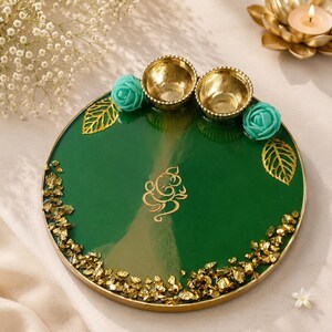 Round green resin pooja thali with a gold-flecked edge, featuring two small gold diyas, turquoise rose accents, gold leaf decorations, and a gold Ganesh emblem etched in the center of the tray.