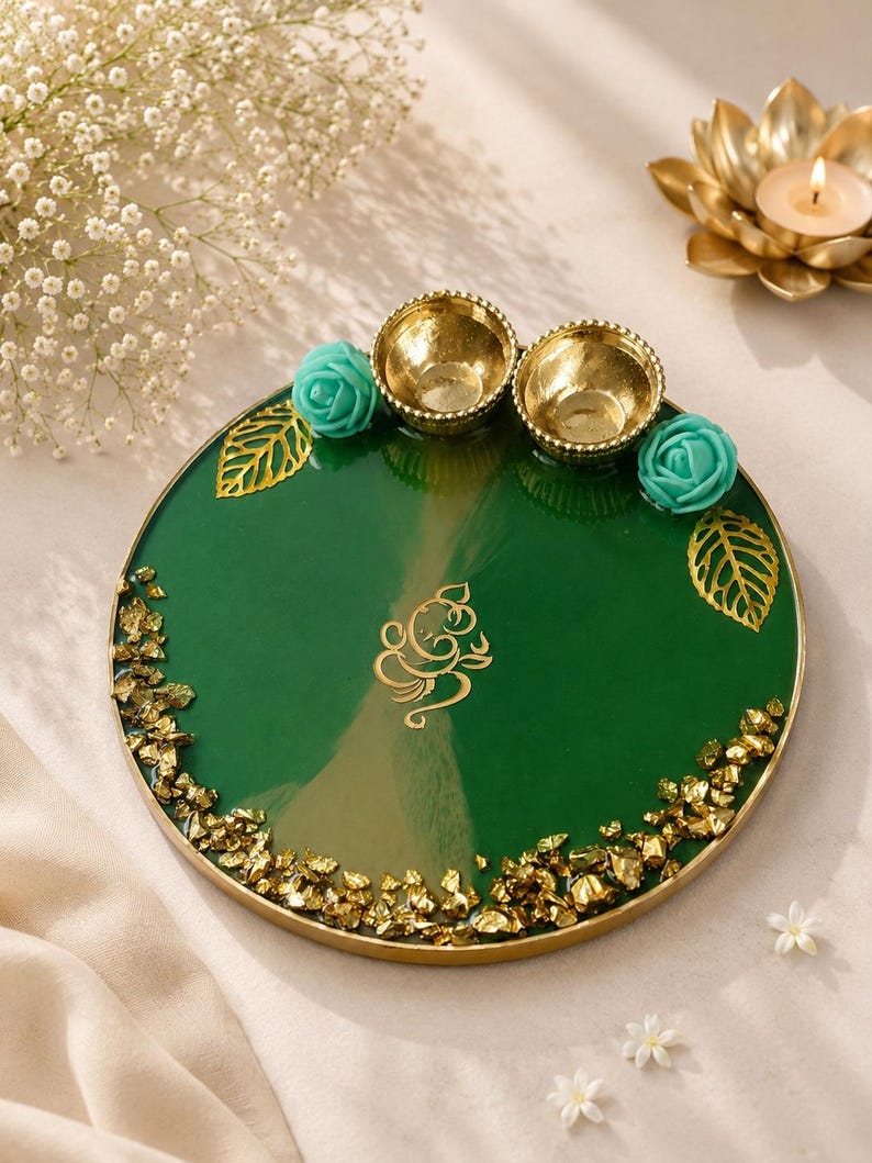 Round green resin pooja thali with a gold-flecked edge, featuring two small gold diyas, turquoise rose accents, gold leaf decorations, and a gold Ganesh emblem etched in the center of the tray.